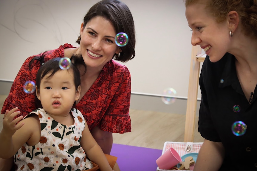 Two women smile at bubbles as a small child plays with them. They are all inside.