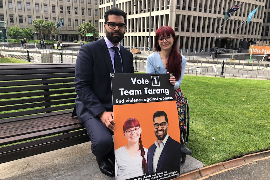 A man in a suit and a red-haired woman with glasses sit on a bench in a park in Melbourne's CBD, holding an election poster.