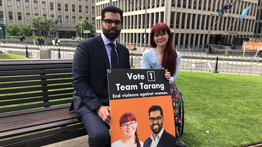 A man in a suit and a red-haired woman with glasses sit on a bench in a park in Melbourne's CBD, holding an election poster.