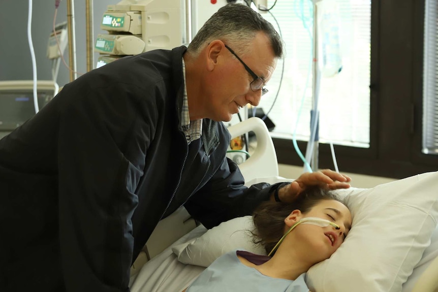 Daniel Lukan touches the head of daughter Grace, who is in a hospital bed