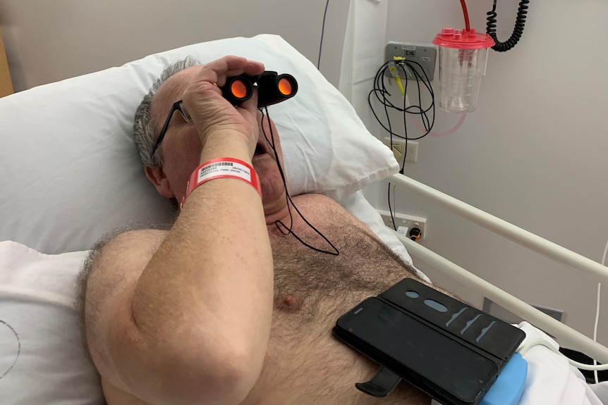 A man laying in a hospital bed using binoculars and looking surprised.