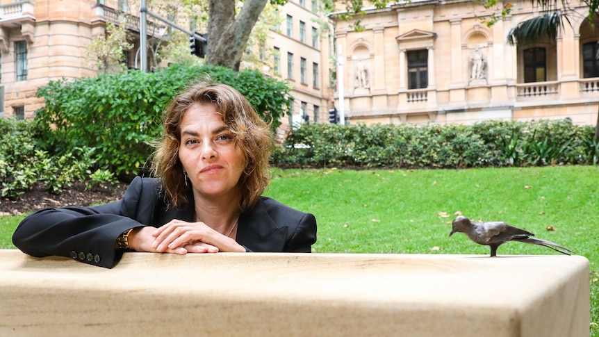 The artist crouches in a Sydney park next to her art work, a small bronze sculpture of a bird mounted onto a sand stone plinth.