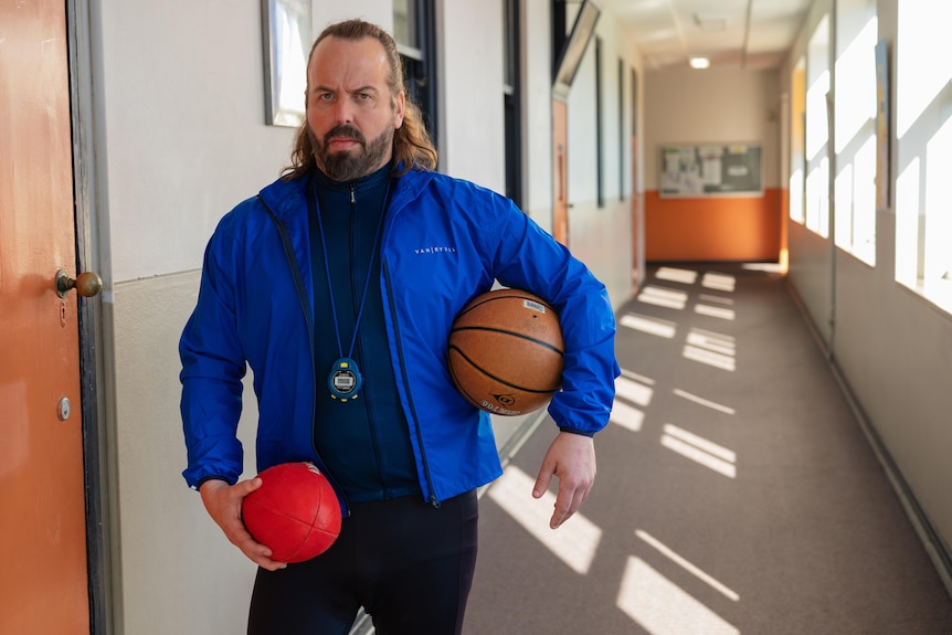 A man in his 40s in a blue jacket roams empty school halls holding a basketball and AFL ball. A whistle is around his neck.