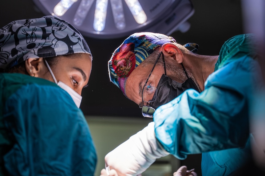 A male doctor and female nurse in surgical masks and green scrubs perform surgery under a bright light