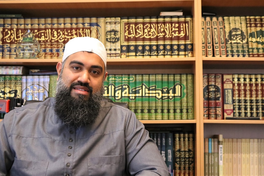 A Muslim Imam wearing his Takiyah skullcap, smiling, in front of a library of Islamic texts.