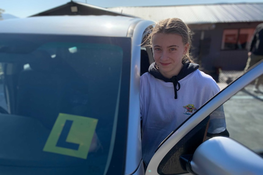 A young woman stands near a car with L plate displayed.