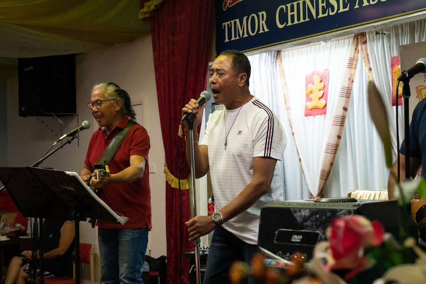Before red curtains, two men on stage sing holding microphones. Behind them is a sign, 'Timor Chinese'.