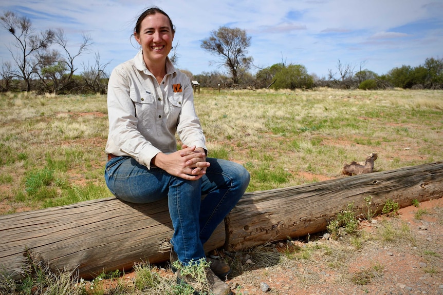 A woman with a brown ponytail, wearing blue jeans and a grey shirt, sits smiling on a log on a grassy plain.