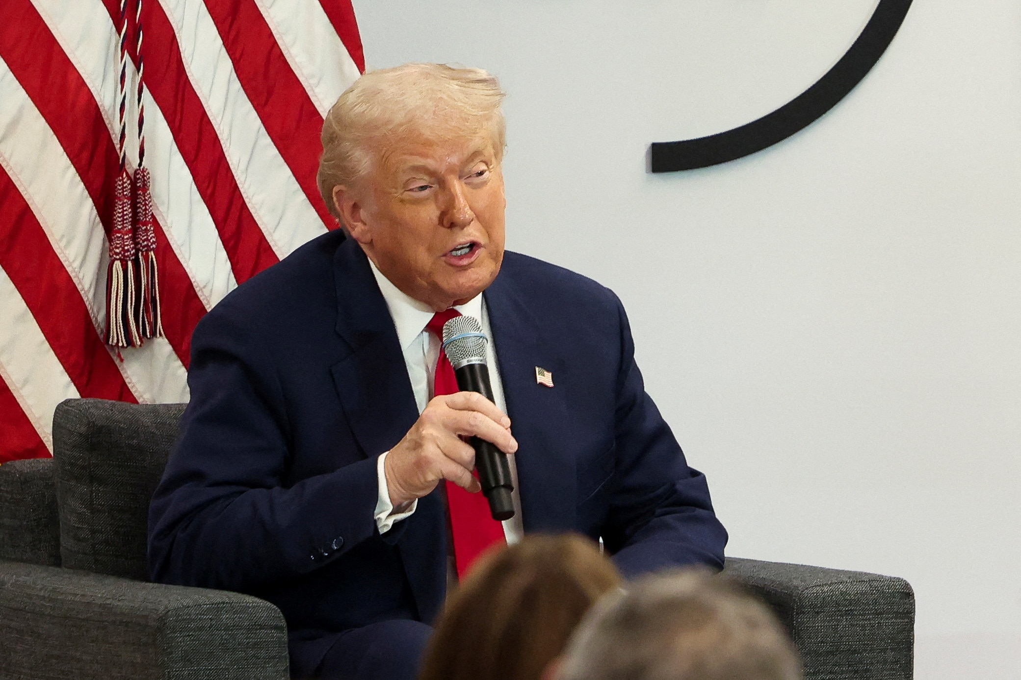 US President Donald Trump sits in a chair holding a microphone in front of a US flag.