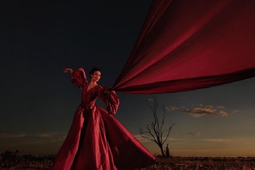 A woman in a red gown stands in the dark in front of an old tree with her scarf blowing in the wind