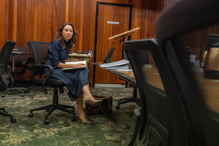 A woman sits at a desk in a court room. She has a notebook on her lap. 
