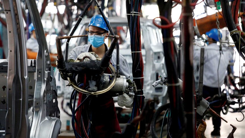 A man works at a car factory. He is wearing a helmet and a mask over his face.