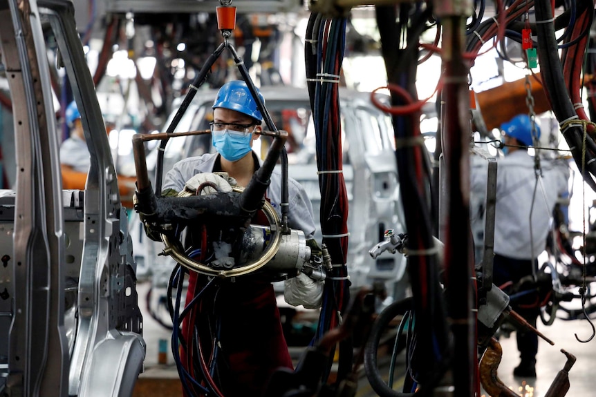 A man works at a car factory. He is wearing a helmet and a mask over his face.