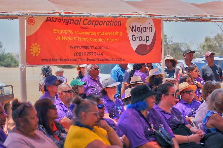 A crowd wearing purple shirts sits behind a banner