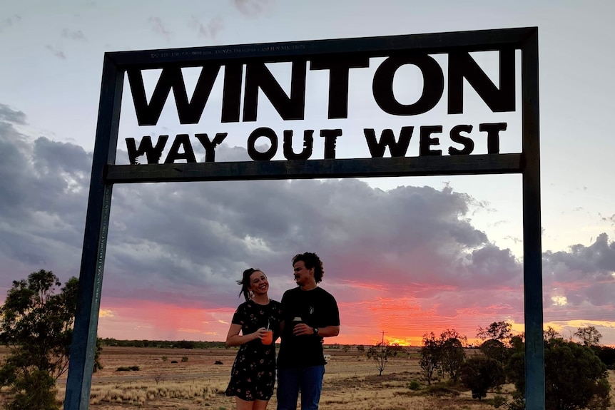 A young couple stand under a sign that reads 'Winton Way Out West' as the sun is setting.