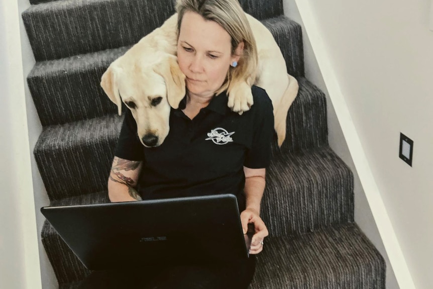 A woman in a dark shirt sits on a set of stairs, looking at a laptop, with a dog resting its head on her shoulder.