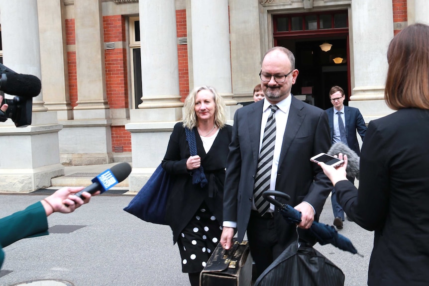 A man and a woman dressed in business attire leave a court building as reporters approach.