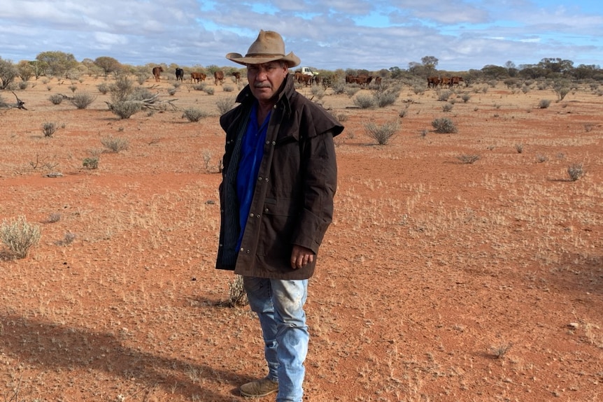 A moustachioed Indigenous man in stockman's getup – Driza-Bone and hat — stands on a red desert plain.