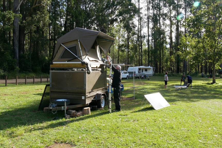Man putting up his ladder on his camper trailer in a green, grassy campsite surrounded by tall trees 
