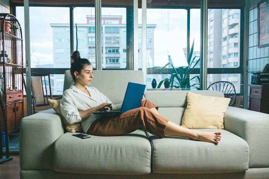 A woman sits on her couch with a laptop on her lap working from home