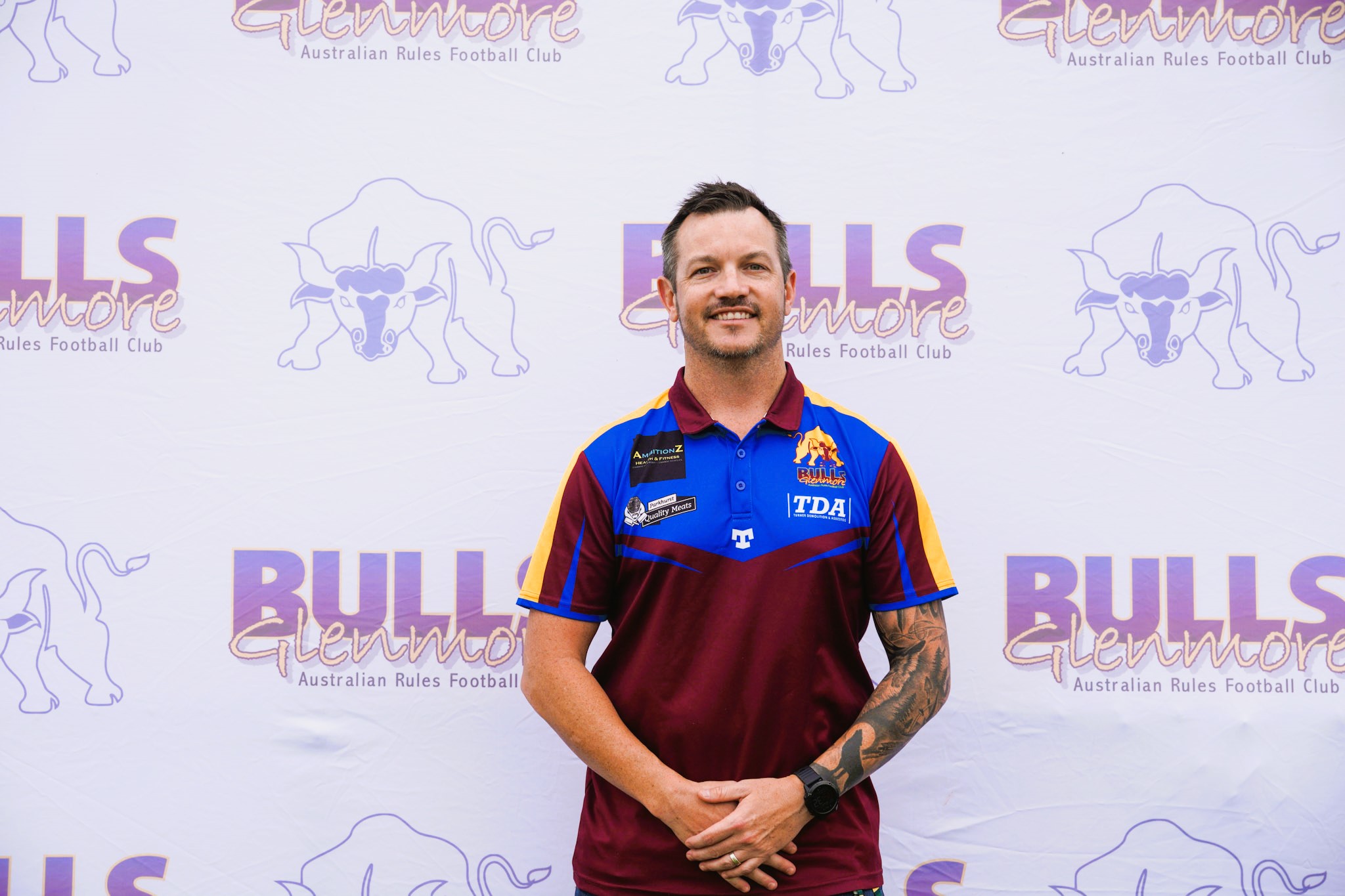 A man wearing a maroon, blue and yellow sports polo standing in front of a team sign.