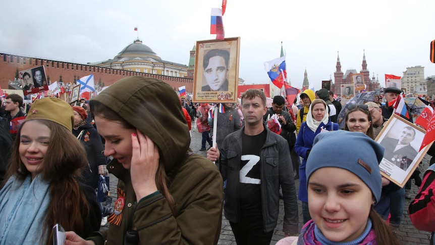 a large crowd in Red Square including a man in a t-shirt with a 'z' on it holding up a picture of a relative that died in WW2