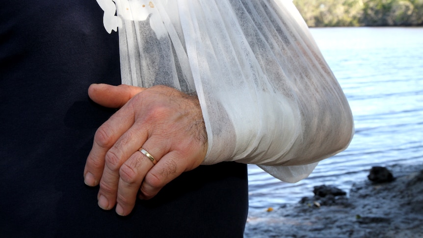A close up shot shows a man's arm with a surgical patch on it, held within a sling. He stands beside a waterway.
