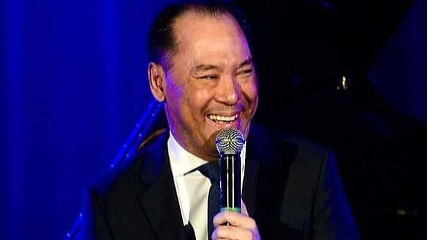 Asian man in balck suit, white shit and black tie talking into a microphone infront of blue background