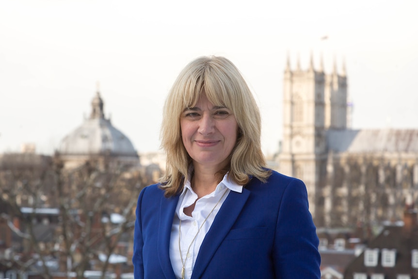 a woman with blond hair and blue blazer stand in focu with westminster palace in the background