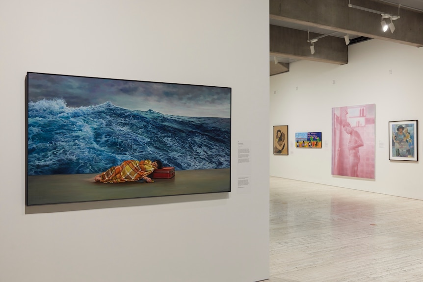 A large oil painting of a woman lying down head on a suitcase wrapped in a blanket, the background is ocean, hanging in gallery