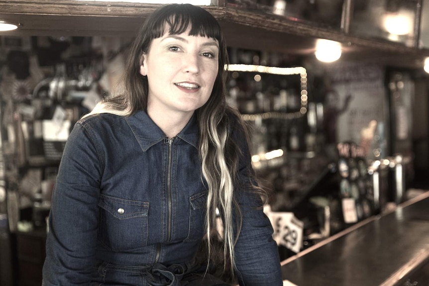 A woman with long hair sits in front of a bar and looks into the camera.