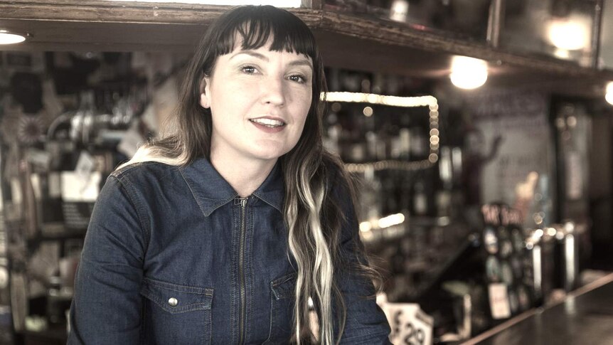 A woman with long hair sits in front of a bar and looks into the camera.