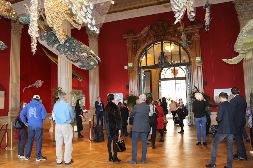 People inside an austere room with sculptures made of netting including fish and a whale hanging from the ceiling.