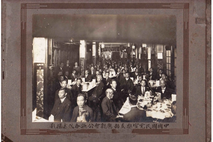 A black and white image shows a crowd of men in suits sitting at round tables in a packed restaurant.