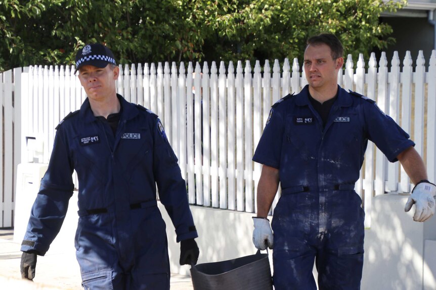 Two police forensic officers walk along a footpath carrying a bucket outside a house.