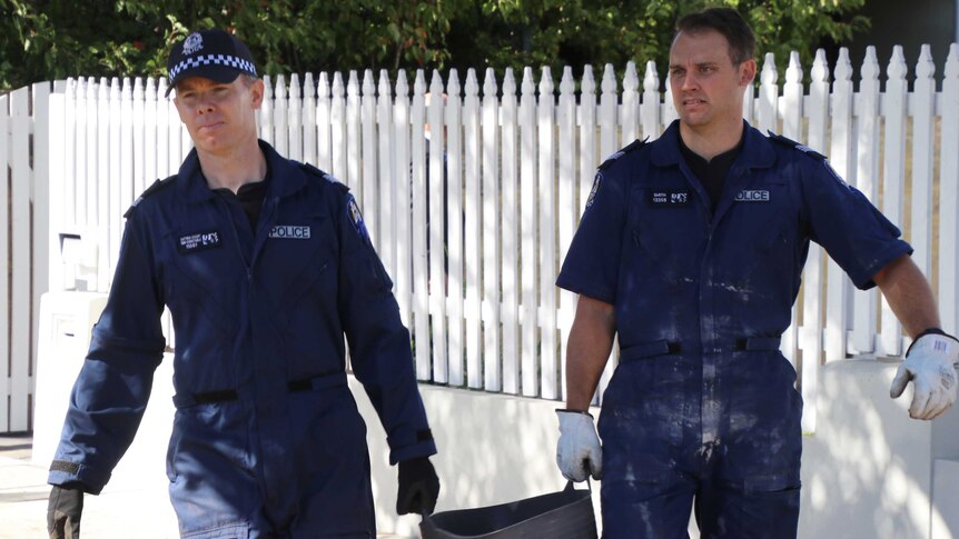 Two police forensic officers walk along a footpath carrying a bucket outside a house.