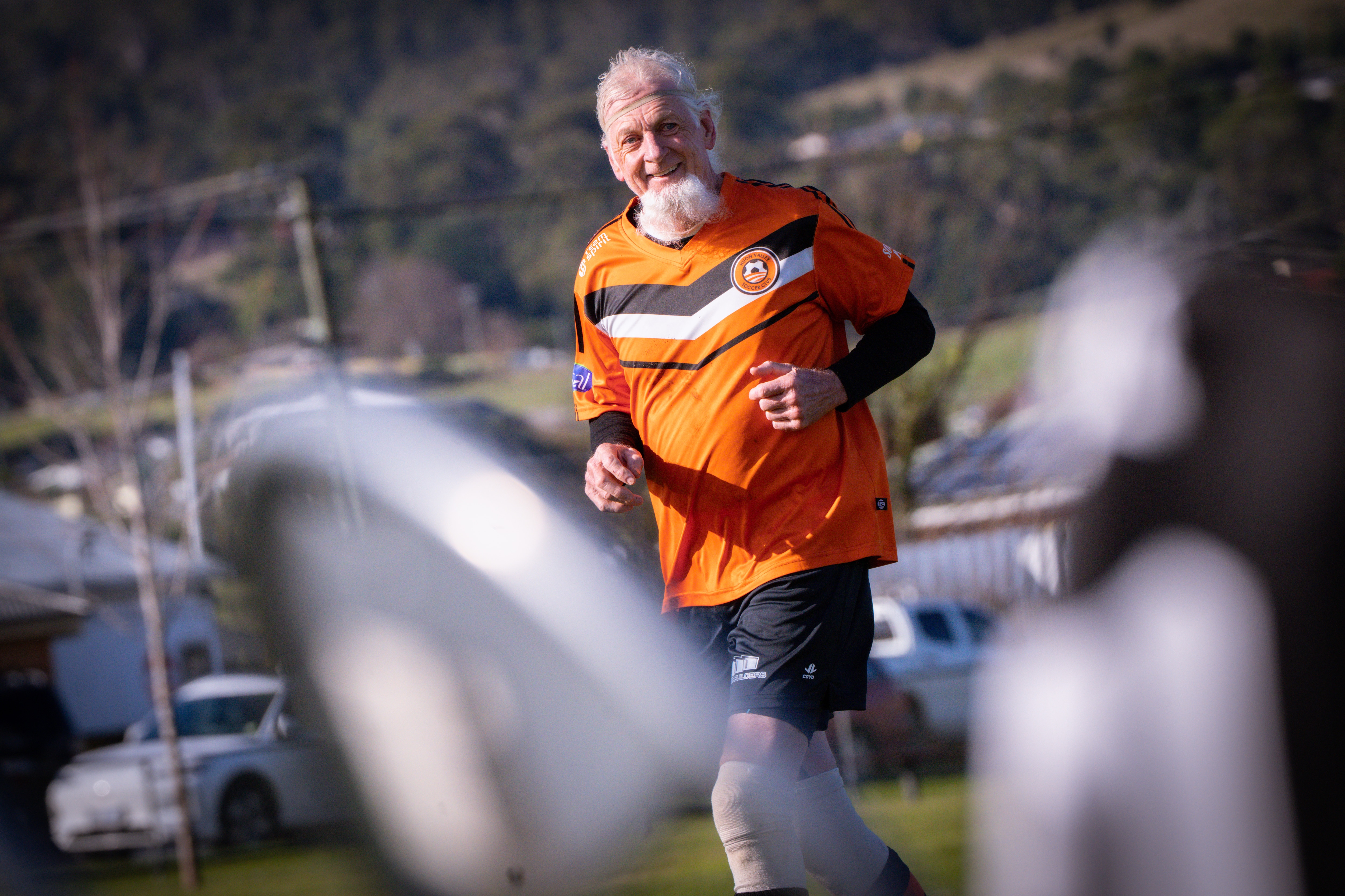 A man with white hair and a long white beard wearing orange soccer colours on a soccer field.