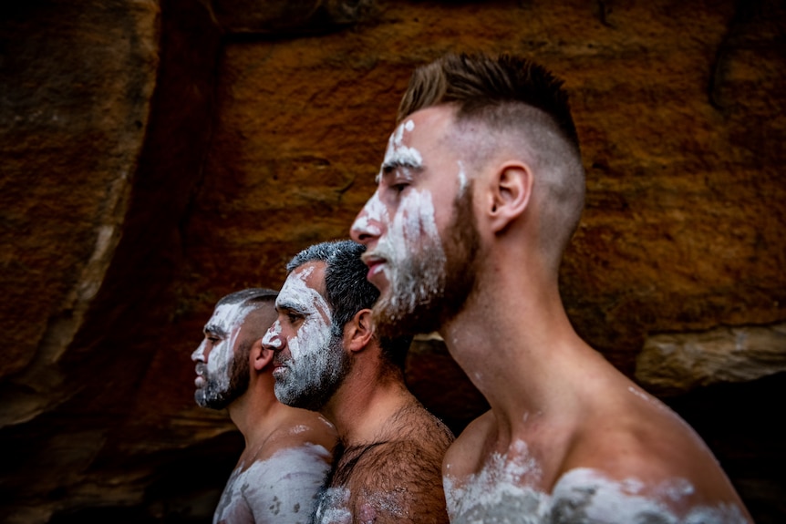 Three men stand in a line with their faces painted.
