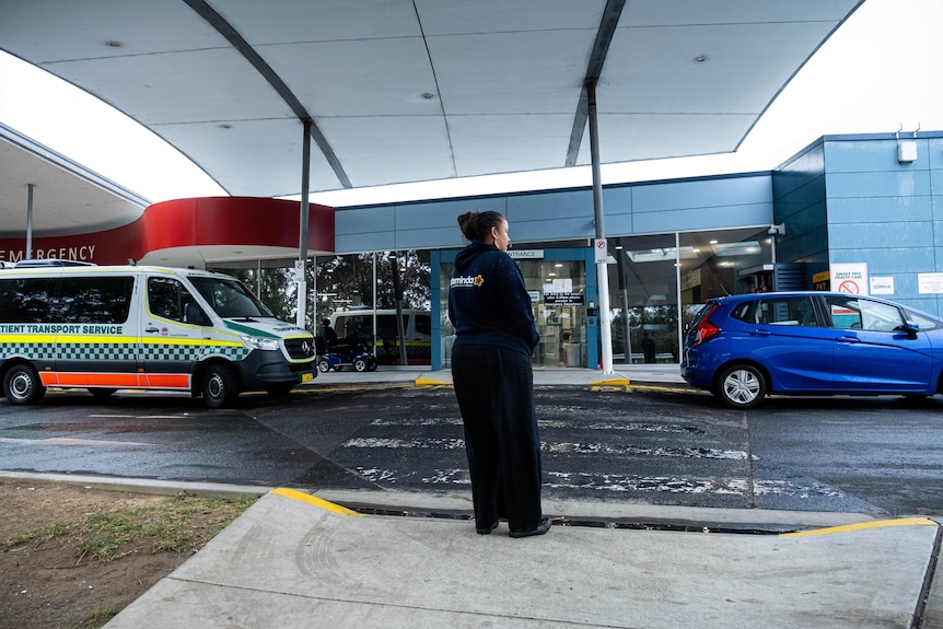 A woman stands in the carpark of a hospital. There’s an ambulance parked to her left. 