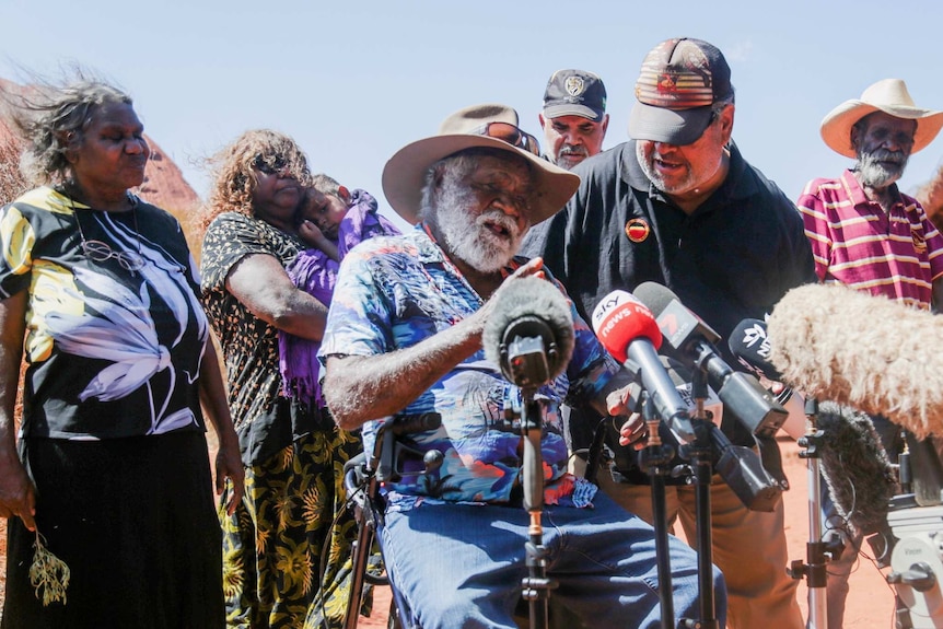 Traditional owner Reggie Uluru sits in front of microphones to speak to the media surrounded by other traditional owners