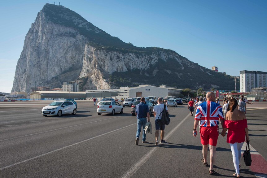 A couple in matching red outfits, a man in a Union Jack jumper, stroll a Gibraltar street with its famed rock in the distance.