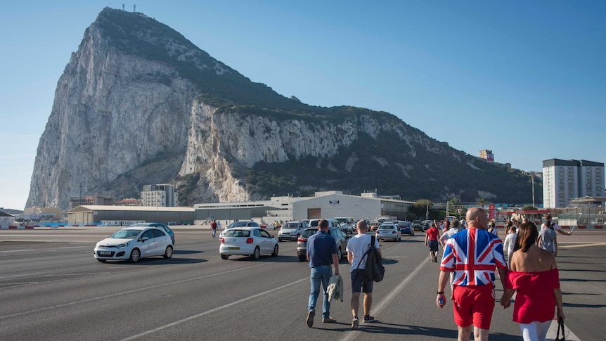 A couple in matching red outfits, a man in a Union Jack jumper, stroll a Gibraltar street with its famed rock in the distance.