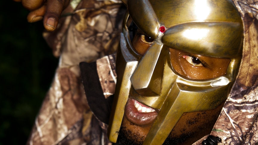 MF DOOM wears a gold mask and glares at the camera