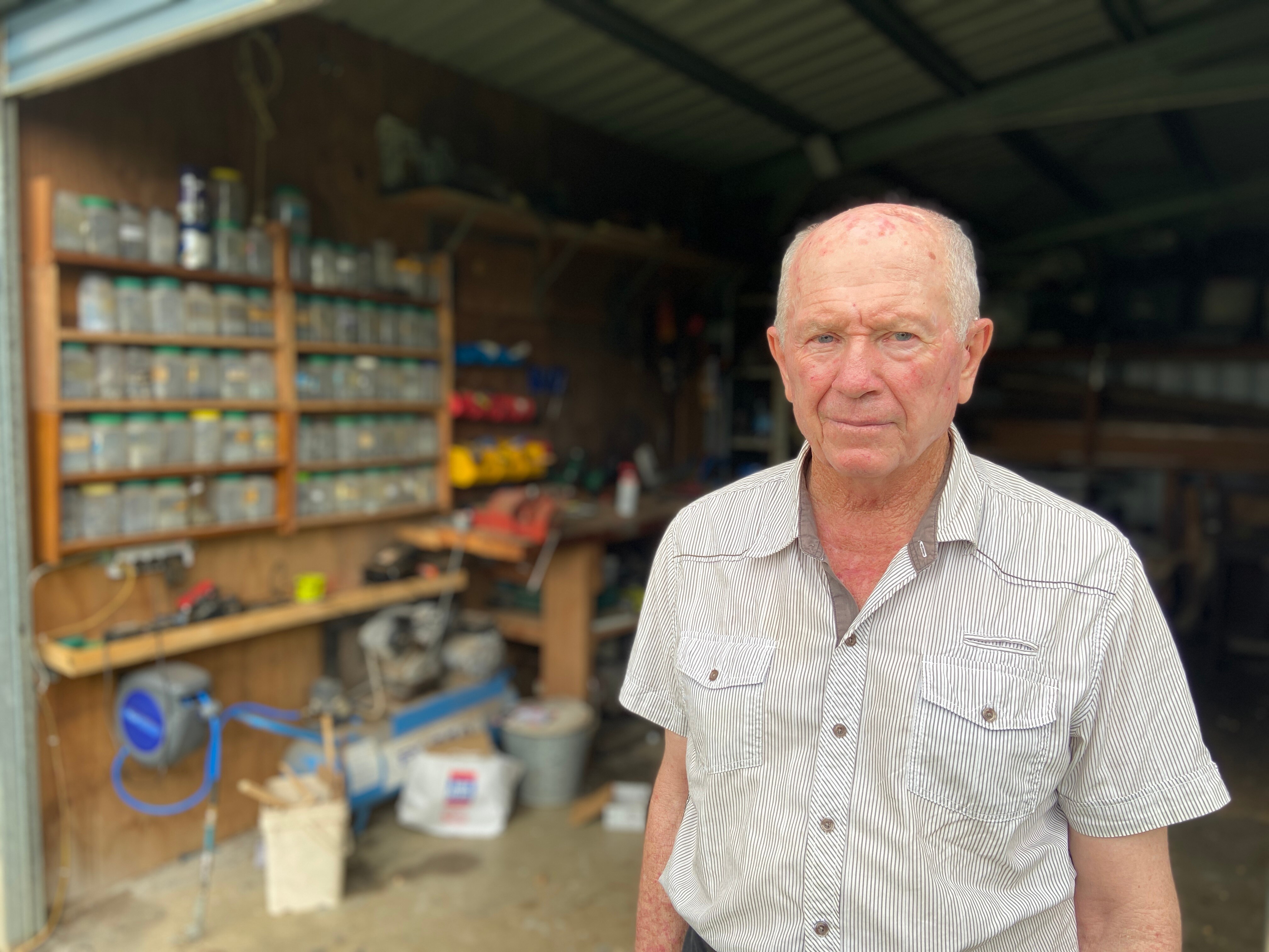 An older man standing in front of an open shed, shelves lined with jars and tools blured in the background.