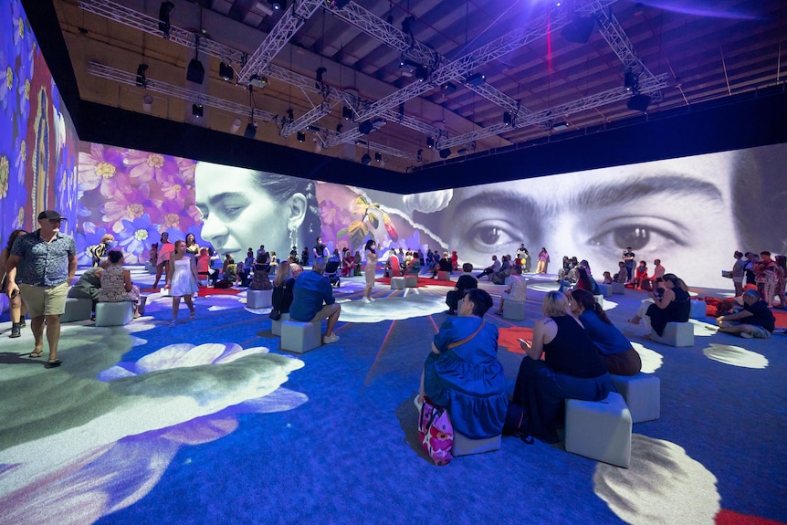 About 50 people sit in an illuminated gallery lined with projection screens featuring montage images of Frida Kahlo and her art.
