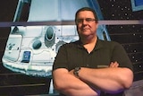 A man stands in front of a space capsule suspended indoors. 