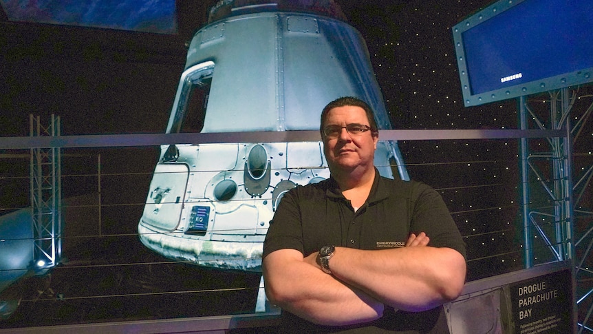 A man stands in front of a space capsule suspended indoors. 