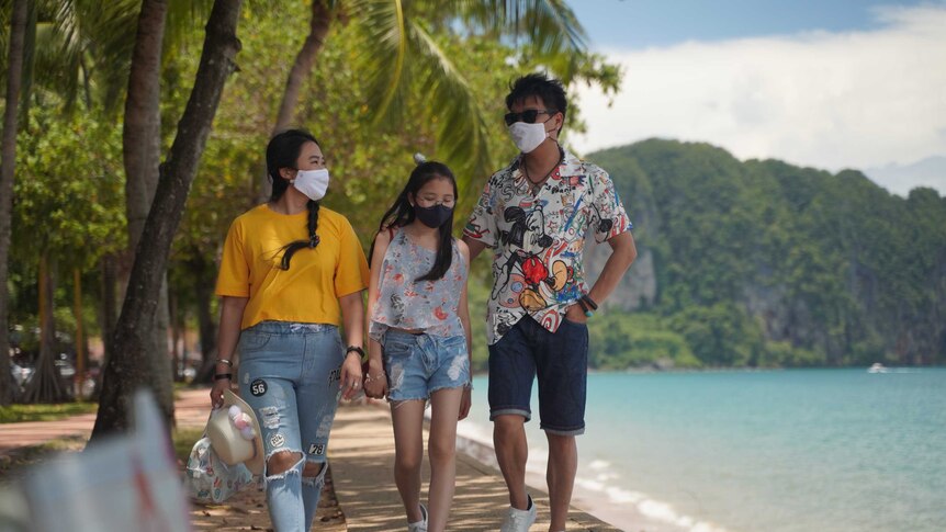 Three people wearing colourful clothes and masks walk along a sidewalk next to a beach with mountains in the background.
