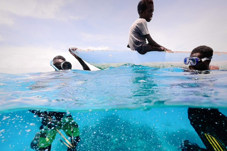 Three boys spearfishing, two in the water while one sits on a boat.