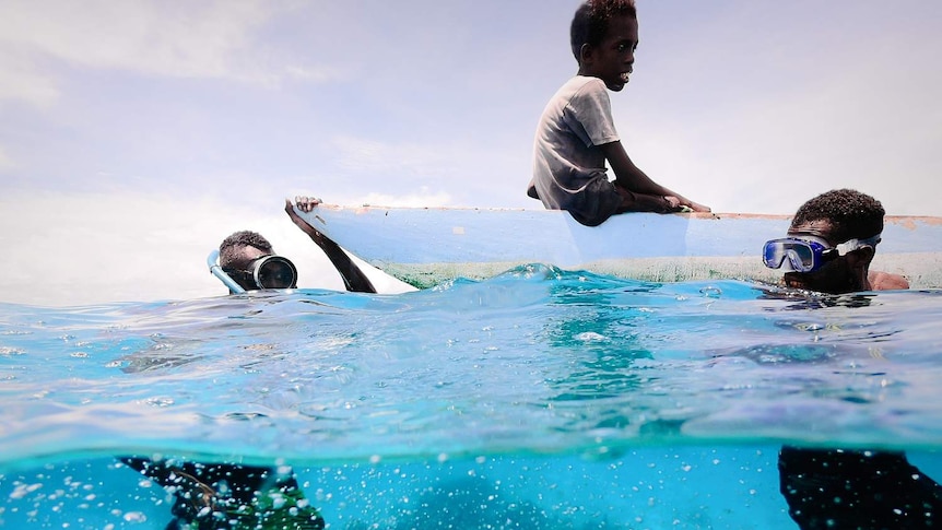 Three boys spearfishing, two in the water while one sits on a boat.